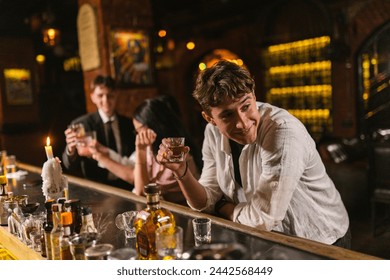 Young guy pulls away from comrades after night party in restaurant. Handsome man drinks alcohol among visitors in modern place