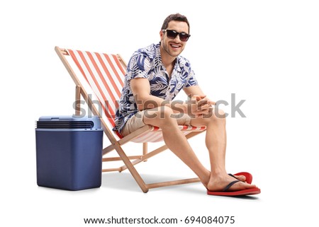 Young guy sitting in a deck chair next to a cooling box and looking at the camera isolated on white background