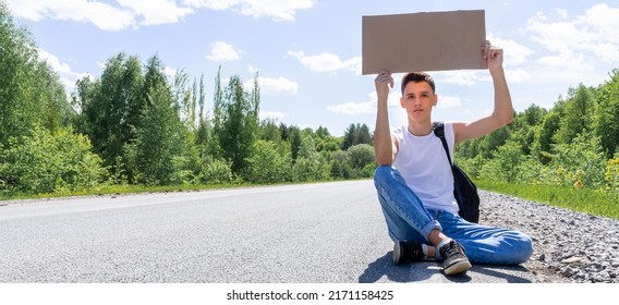 A young guy is sitting on the road with a sign in his hands, hitchhiking around the country. A man is trying to catch a passing car for a trip. A teenager with a backpack went on vacation hitchhiking