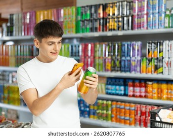 Young guy stands in drinks section of supermarket and uses his phone to find out information about drink in metal can in his hand