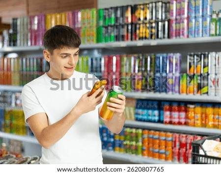 Young guy stands in drinks section of supermarket and uses his phone to find out information about drink in metal can in his hand