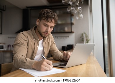 Young Guy Student Prepare Research Essay At Modern Kitchen Surf Internet Use Laptop Write Up Information To Paper Report By Hand. Focused Man Freelancer Work From Home Make List Of Data From Pc Screen