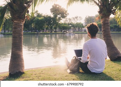 Young Guy Study While Sitting On The Grass By The Water In A Park. Warm Tones In Sunset. Copy Space. Study Or Freelance Working Concept.