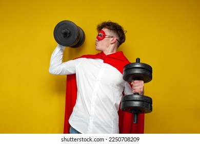 young guy in a superman costume lifts heavy dumbbells and does sports on a yellow background, a super hero with super strength and superpowers