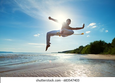 A young guy trains capoeira on the beach.  - concept about people, lifestyle and sport. A boy performs martial the kick in the jump. The silhouette against the sunlight