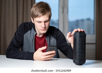 Young Guy Turns On The Bluetooth Speaker. He Controls The Portable Speaker Through His Smartphone. Portrait Of A Student With A Portable Column. Concept - Intelligent Voice-activated Speaker.