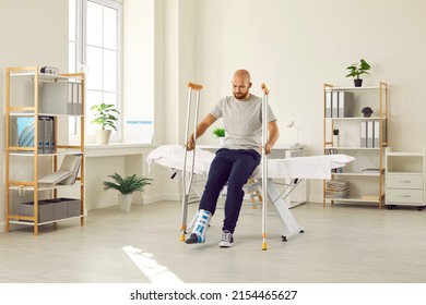 Young Guy Undergoing Rehabilitation After Foot Injury. Male Patient Wearing Support Brace And Fracture Fixator On Broken Leg Sitting On Couch At Hospital And Trying To Stand Up With Help Of Crutches