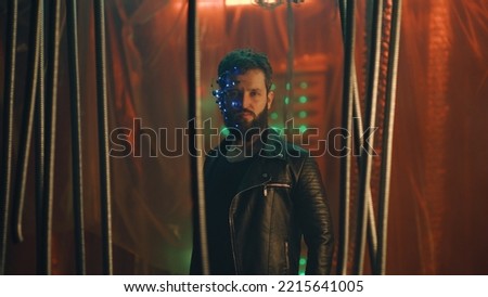 Young guy wearing a headset with LED lights stands behind the black hanging rods and looks at the camera. Cyberpunk and futuristic concept. Neon lights in the background.