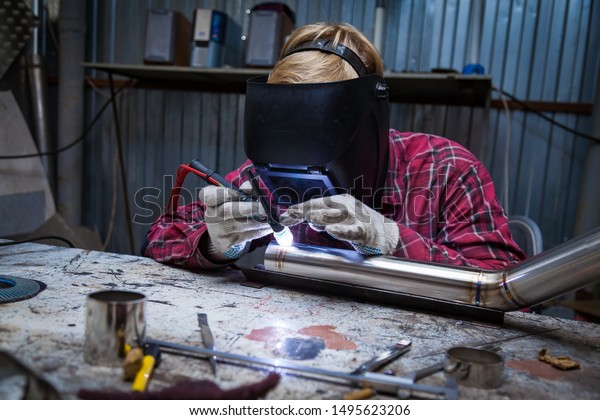 Young guy welder in a\
checkered red shirt welds a stainless steel pipe using agronomic\
welding to protect his eyes with a mask in an iron workshop. Modern\
welding methods.