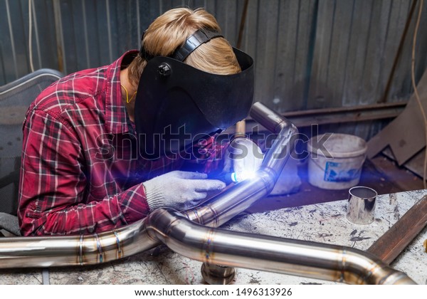 Young\
guy welder in a checkered red shirt welds a stainless steel pipe\
for car exhaust using agronomic welding to protect his eyes with a\
mask in an iron workshop. Modern welding\
methods.