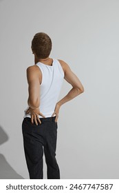 Young guy in white singlet and black pants posing and posing isolated on grey studio background. Back view. Muscular body. Concept of male beauty, body care, fitness, sport, health, fashion
