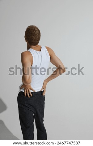 Young guy in white singlet and black pants posing and posing isolated on grey studio background. Back view. Muscular body. Concept of male beauty, body care, fitness, sport, health, fashion