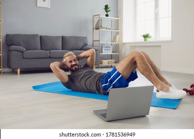 Young Guy Working Out To Online Video Tutorial On Laptop At Home. Sporty Man Doing Abs Exercises On Yoga Mat Indoors