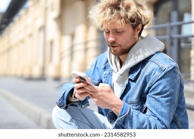 Young guy writes a message in the phone while sitting on the street on a sunny day.