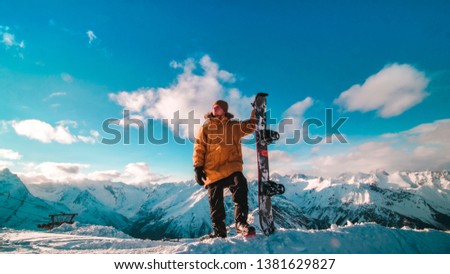 A young guy in a yellow down jacket with a snowboard in his hands stands on the top of the mountain. Snowboarder stands on the background of a beautiful view of the snow-capped mountains.
