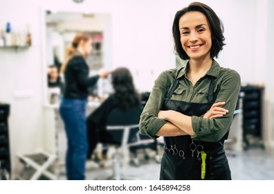 Young hairdresser. Portrait of professional hairstylist looking in the camera and smiling with folded arms, standing near client who is having her hair done.