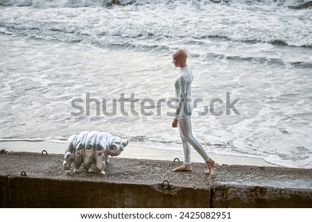 Young hairless girl with alopecia in white futuristic suit walking on concrete fence with toy tardigrade on sea background, bald pretty girl symbolizes courage and acceptance of unique appearance