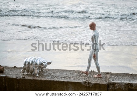 Young hairless girl with alopecia in white futuristic suit walking on concrete fence with toy tardigrade on sea background, bald pretty girl symbolizes courage and acceptance of unique appearance