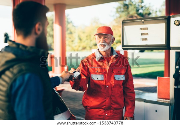 Young handsome adult man\
together with senior worker standing on gas station and fueling\
car.