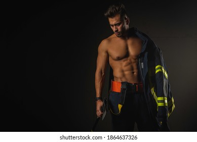 young handsome adult, muscular firefighter in uniform holding ax of fire equipment in his hands, pensive, isolated on dark background. Low key. Protection concept. there is a place for an inscription,