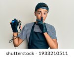 Young handsome african american man tattoo artist wearing professional uniform and gloves holding tattooer machine asking to be quiet with finger on lips. silence and secret concept. 