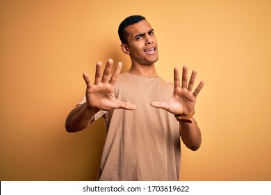 Young handsome african american man wearing casual t-shirt standing over yellow background afraid and terrified with fear expression stop gesture with hands, shouting in shock. Panic concept.