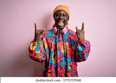 Young Handsome African American Man Wearing Colorful Coat And Cap Over Pink Background Shouting With Crazy Expression Doing Rock Symbol With Hands Up. Music Star. Heavy Concept.