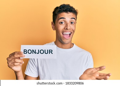 Young handsome african american man holding bonjour french greeting word celebrating achievement with happy smile and winner expression with raised hand 
