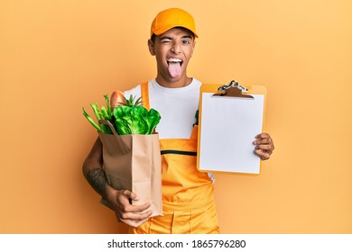 Young handsome african american man wearing courier uniform with groceries from supermarket and clipboard sticking tongue out happy with funny expression. 