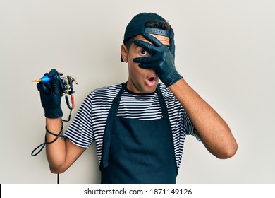 Young Handsome African American Man Tattoo Artist Wearing Professional Uniform And Gloves Holding Tattooer Machine Peeking In Shock Covering Face And Eyes With Hand, Looking Through Fingers