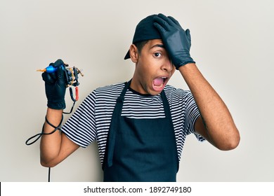 Young Handsome African American Man Tattoo Artist Wearing Professional Uniform And Gloves Holding Tattooer Machine Surprised With Hand On Head For Mistake, Remember Error. Forgot, Bad Memory Concept. 