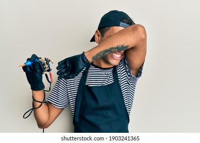Young Handsome African American Man Tattoo Artist Wearing Professional Uniform And Gloves Holding Tattooer Machine Smiling Cheerful Playing Peek A Boo With Hands Showing Face. Surprised And Exited 