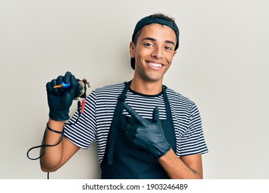 Young Handsome African American Man Tattoo Artist Wearing Professional Uniform And Gloves Holding Tattooer Machine Cheerful With A Smile On Face Pointing With Hand And Finger Up To The Side Happy