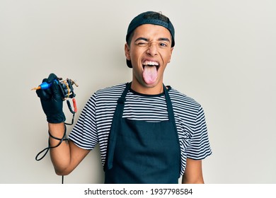 Young Handsome African American Man Tattoo Artist Wearing Professional Uniform And Gloves Holding Tattooer Machine Sticking Tongue Out Happy With Funny Expression. Emotion Concept. 