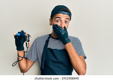 Young Handsome African American Man Tattoo Artist Wearing Professional Uniform And Gloves Holding Tattooer Machine Shocked Covering Mouth With Hands For Mistake. Secret Concept. 