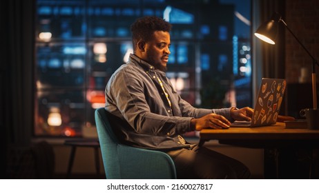 Young Handsome African American Man Working From Home On Laptop Computer In Stylish Loft Apartment At Night. Creative Male Checking Social Media, Browsing Internet. Urban City View From Big Window.