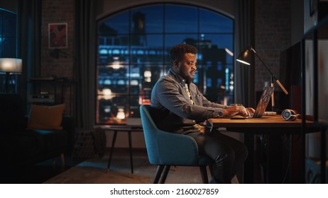 Young Handsome African American Man Working From Home On Laptop Computer In Stylish Loft Apartment At Night. Creative Male Checking Social Media, Browsing Internet. Urban City View From Big Window.