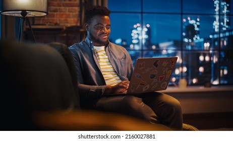 Young Handsome African American Man Working From Home On Laptop Computer In Stylish Loft Apartment At Night. Creative Male Checking Social Media, Browsing Internet. Urban City View From Big Window.