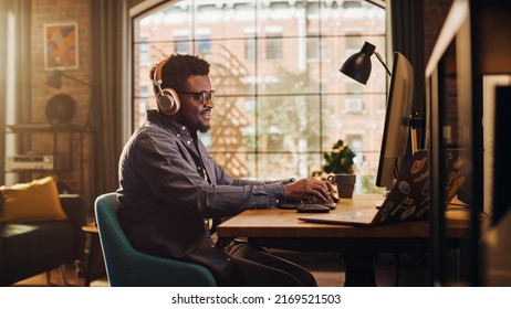 Young Handsome African American Man Working from Home on Desktop Computer in Sunny Stylish Loft Apartment. Creative Male Checking Social Media, Browsing Internet. Urban City View from Big Window.