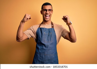 Young handsome african american shopkeeper man wearing apron over yellow background celebrating surprised and amazed for success with arms raised and open eyes. Winner concept.