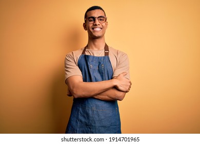 Young handsome african american shopkeeper man wearing apron over yellow background happy face smiling with crossed arms looking at the camera. Positive person.