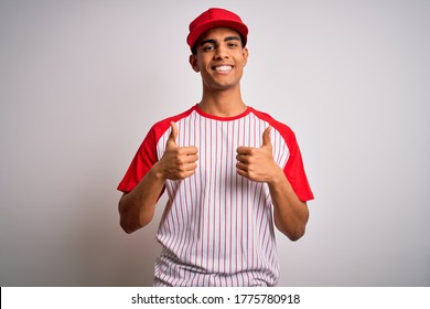 Young handsome african american sportsman wearing striped baseball t-shirt and cap success sign doing positive gesture with hand, thumbs up smiling and happy. Cheerful expression and winner gesture.