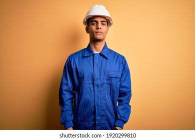 Young handsome african american worker man wearing blue uniform and security helmet with serious expression on face. Simple and natural looking at the camera.