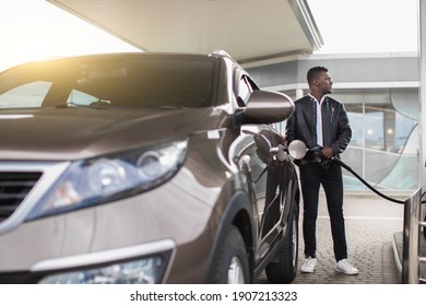 Young handsome African bearded guy in stylish casual outfit refueling his luxury car and looking away while standing on self service petrol station