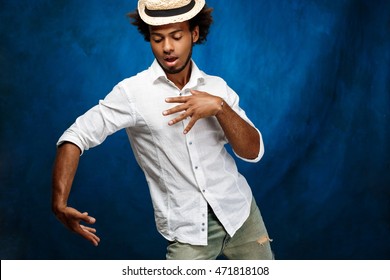 Young Handsome African Man In Hat Dancing Over Blue Background.