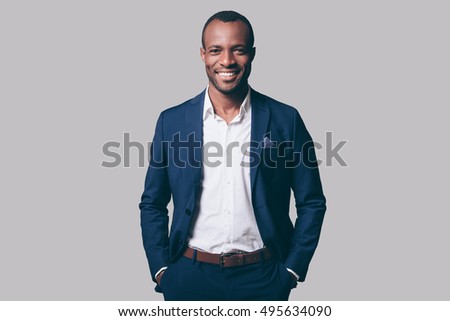 Young and handsome. Handsome young African man in smart casual jacket holding hands in pockets and smiling while standing against grey background