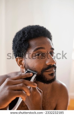 Young handsome African man taking care of his beard, trimming it with an electrical trimmer, beard care routine.