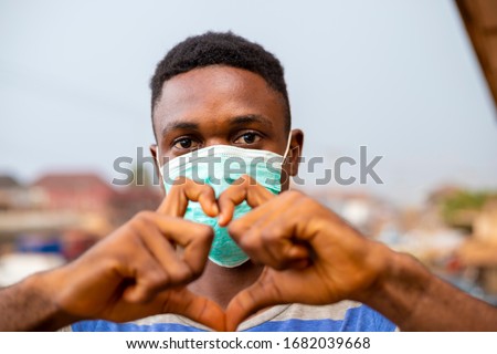 young handsome african nigerian man wore face mask preventing, prevent, prevented himself from the outbreak in his society and did a love sign.