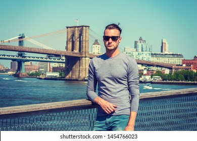 Young Handsome American Man Traveling In New York City, Wearing Gray, Long Sleeve T Shirt, Sunglasses, Standing By East River, Under Sun, Looking Forward. Manhattan, Brooklyn Bridges On Background.
