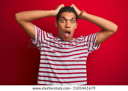 Young handsome arab man wearing striped t-shirt over isolated red background Crazy and scared with hands on head, afraid and surprised of shock with open mouth